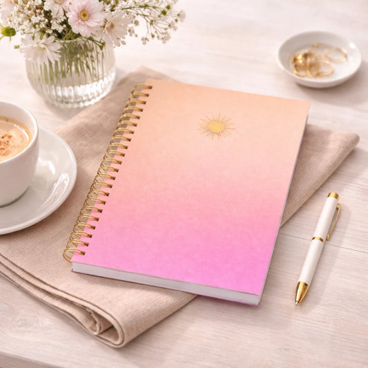spiralblock med gradient i rosa och persikofärgade toner och solsymbol i guld, som ligger på ett bord.